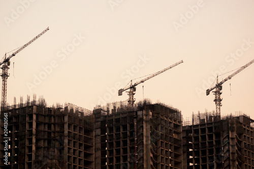 3 under construction buildings with the concrete and iron skeleton and construction cranes at the top shot against the orange sky of dusk. A common site in delhi, noida, jaipur, gurgaon, lucknow