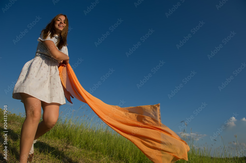 Obraz premium BEAUTIFUL BLONDE GIRL LAUGHS AND DANCES OUTDOORS WITH AN ORANGE SCARF IN HER HANDS