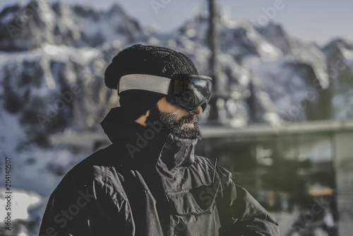 Close-up portrait of bearded handsome snowboarded in sunglass mask, at the ski resort on the background of mountains and blue sky.Blurred background.Horizontal. © KleverLevel