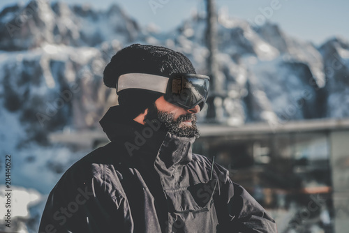 Portrait of bearded handsome snowboarded in sunglass mask, at the ski resort on the background of mountains and blue sky.Blurred background.Horizontal.Color mask © KleverLevel
