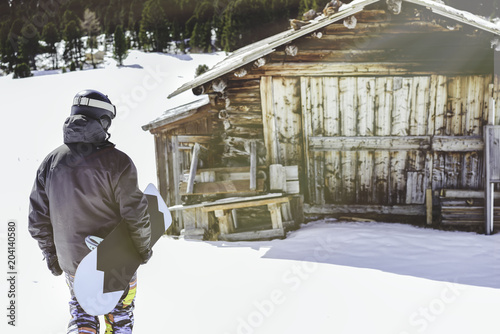 Snowboarder wearing black jacket, mask with snowboard close authentic wooden house.Man walking with snow board desk. Horizontal photo