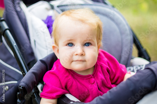 Portrait of a little funny child girl blond with blue eyes sitting in a baby stroller in the summer for greens. Trinasport for a child and transportation of children one year from birth