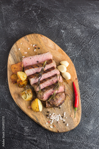 Grilled beef steak with spices, garlic, rosemary and chili pepper on cutting board. Top view.  Stone background