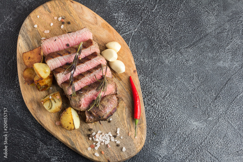 Grilled beef steak with spices, garlic, rosemary and chili pepper on cutting board. Top view. Copy space. Stone background
