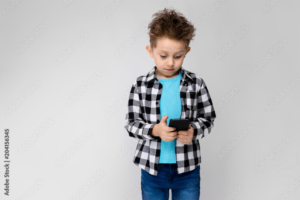 A handsome boy in a plaid shirt, blue shirt and jeans stands on a gray background. The boy is holding a phone A handsome boy in a plaid shirt, blue shirt and jeans stands on a gray background. The boy is holding a phone