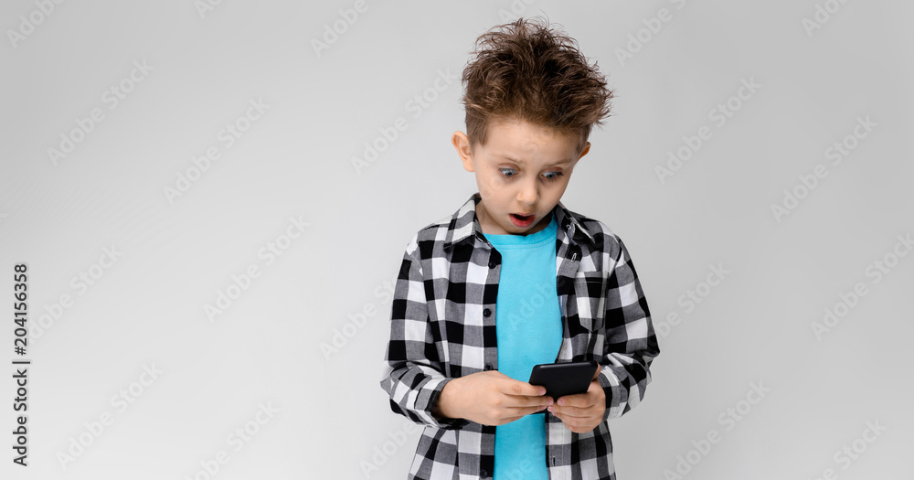 A handsome boy in a plaid shirt, blue shirt and jeans stands on a gray background. The boy is holding a phone A handsome boy in a plaid shirt, blue shirt and jeans stands on a gray background. The boy is holding a phone