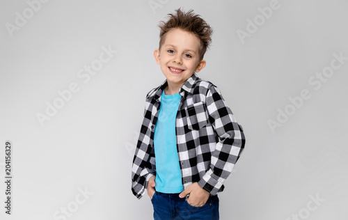 A handsome boy in a plaid shirt, blue shirt and jeans stands on a gray background. The boy smiles and puts his hands into the pockets of his jeans