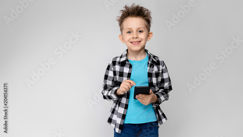 A handsome boy in a plaid shirt, blue shirt and jeans stands on a gray background. The boy is holding a phone