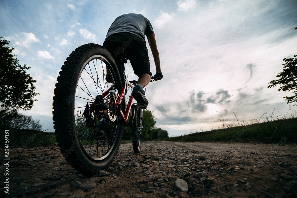 Obraz premium Young man in helmet riding a bicycle along country road on sunset sky background. Bicycle sports, traveling, healthy lifestyle and activity.