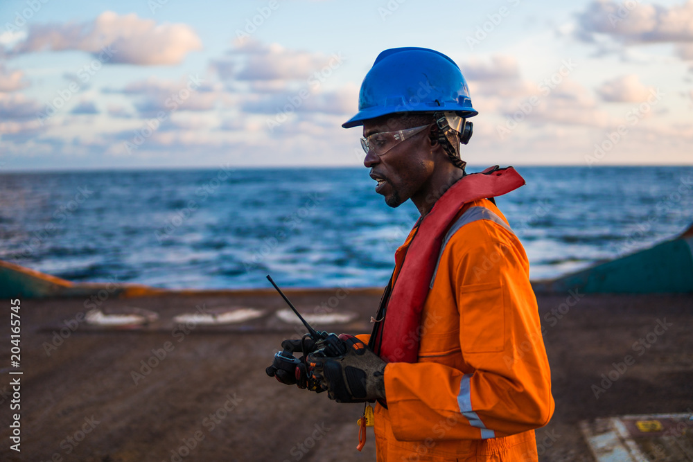 Tired Seaman AB or Bosun on deck of vessel or ship , wearing PPE personal protective equipment - helmet, coverall, lifejacket, goggles. Safety at sea. He has VHF walkie talkie radio in hand Tired Seaman AB or Bosun on deck of vessel or ship , wearing PPE personal protective equipment - helmet, coverall, lifejacket, goggles. Safety at sea. He has VHF walkie talkie radio in hand