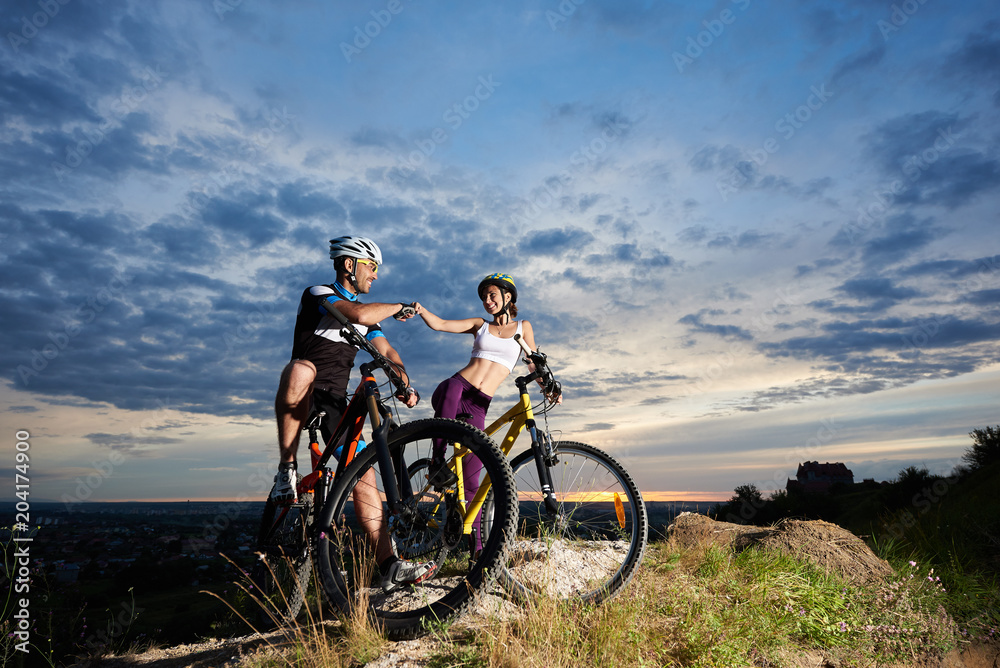 Woman and man on bicycles looking at each other with a smile under the blue sky at sunset. Beautiful scenery on the city opens from the top of the cliff