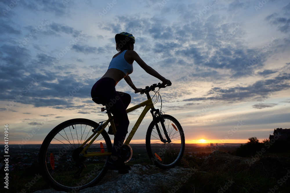 Side view of young lady cycling with an evening landscape. Girl in helmet enjoys sunset on the top of mountain against backdrop of city in distance under fairytale sky with clouds and bright sun