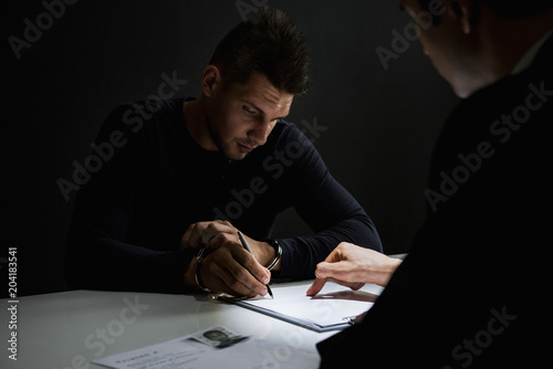 Criminal man with handcuffs signing document in interrogation room photo