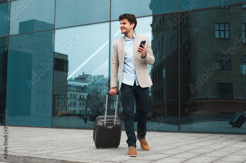 Do your work faster! Full length photo of the cheerful good-looking man going to the business trip and walking with his suitcase. photo