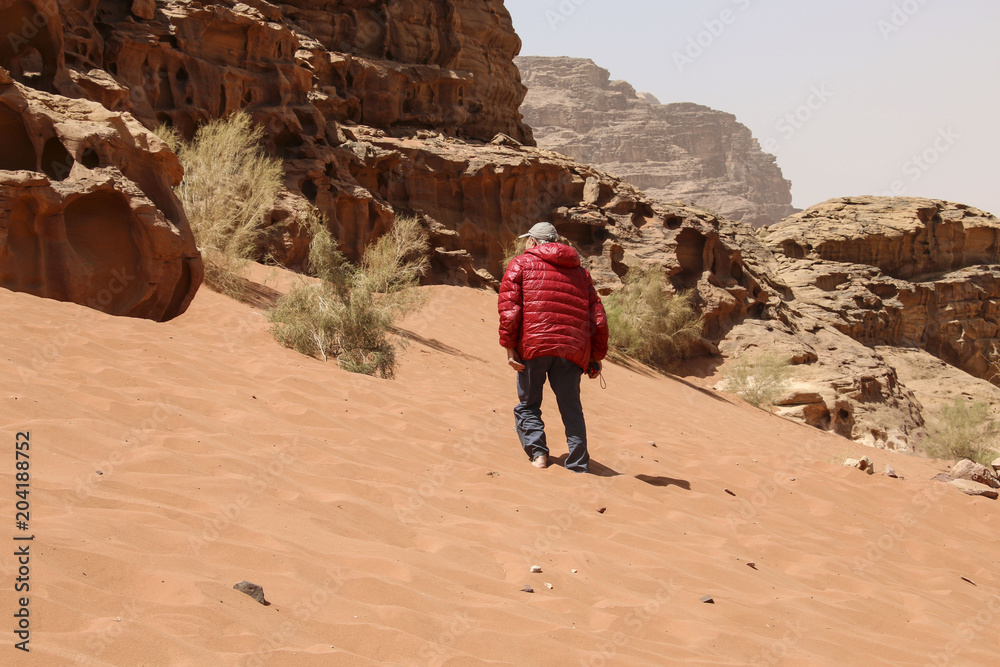 Man in a red jacket rises to a dune of red sand of the canyon of Wadi Rum desert in Jordan.