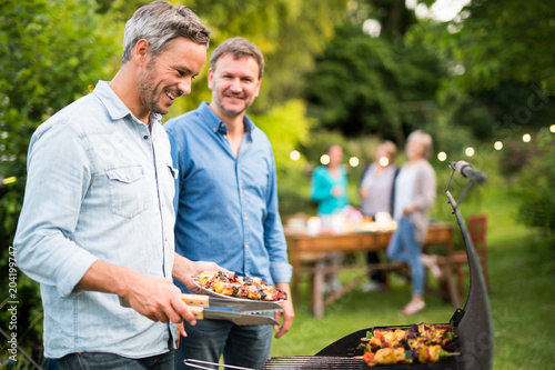 in a summer evening,  two men  in their forties prepares a barbecue for  friends gathered around a table in the garden photo