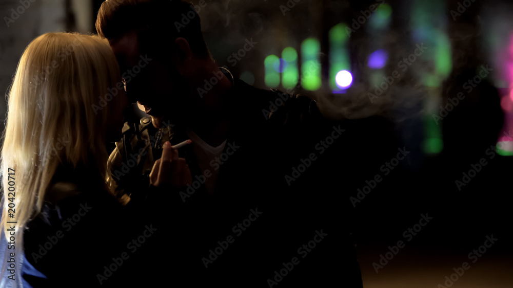 Blonde woman smoking, handsome man embracing her, flirting near nightclub