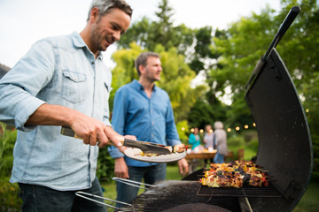 in a summer evening,  two men  in their forties prepares a barbecue for  friends gathered around a table in the garden