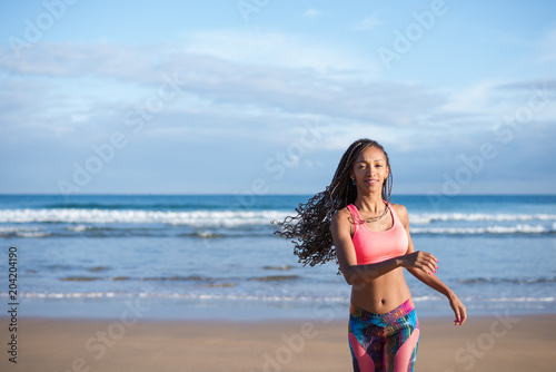 Sporty woman with dreadlocks running at the beach against the sea. Fitness summer lifestyle.