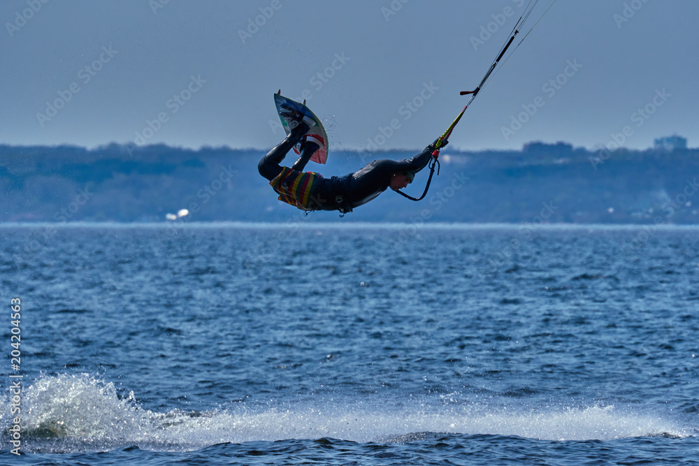 Naklejka premium A male kiteboarder rides on a board on a large river. He performs various exercises while moving on water. Splashes of water scatter in different directions. The sun's rays shine in the water. 
