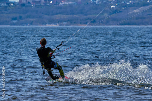 A male kiteboarder rides on a board on a large river. He performs various exercises while moving on water. Splashes of water scatter in different directions. The sun's rays shine in the water.  © afefelov68