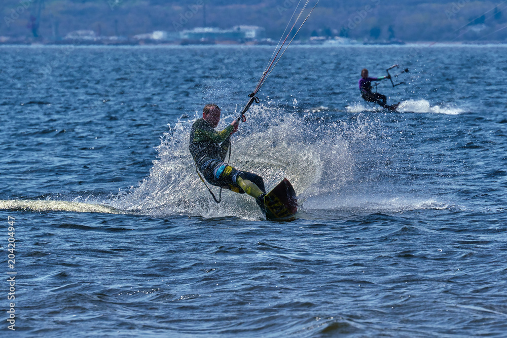 Obraz premium A male kiteboarder rides on a board on a large river. He performs various exercises while moving on water. Splashes of water scatter in different directions. The sun's rays shine in the water. 