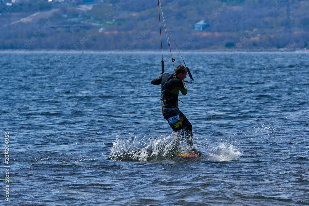 Obraz premium A male kiteboarder rides on a board on a large river. He performs various exercises while moving on water. Splashes of water scatter in different directions. The sun's rays shine in the water. 