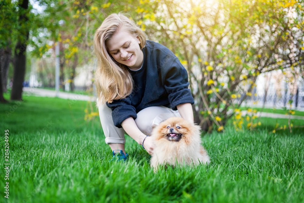 Cute teen girl with blond hair playing with her Pomeranian puppy on green grass in the park Cute teen girl with blond hair playing with her Pomeranian puppy on green grass in the park