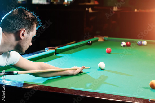 A man in a white T-shirt is playing billiards, a dark background. Pleasant pastime, entertainment, leisure, family holidays, games.