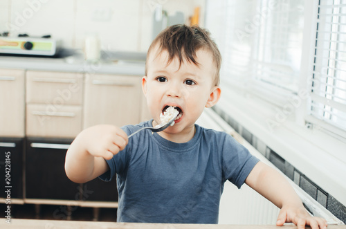 The little boy in the kitchen eagerly eating rice with a spoon independently photo