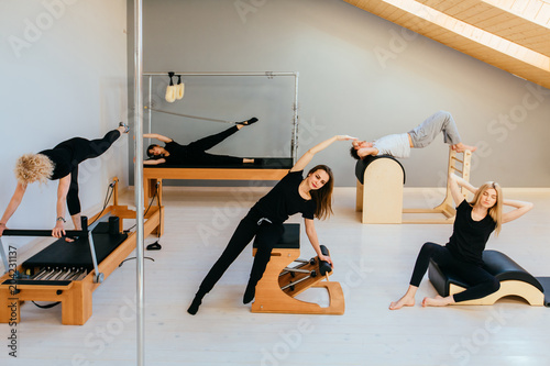 Carta da parati Group of fшму people doing pilates exercises with chair, barrel, spine corrector, reformer, cadillac - different equipment in modern eco studio interior