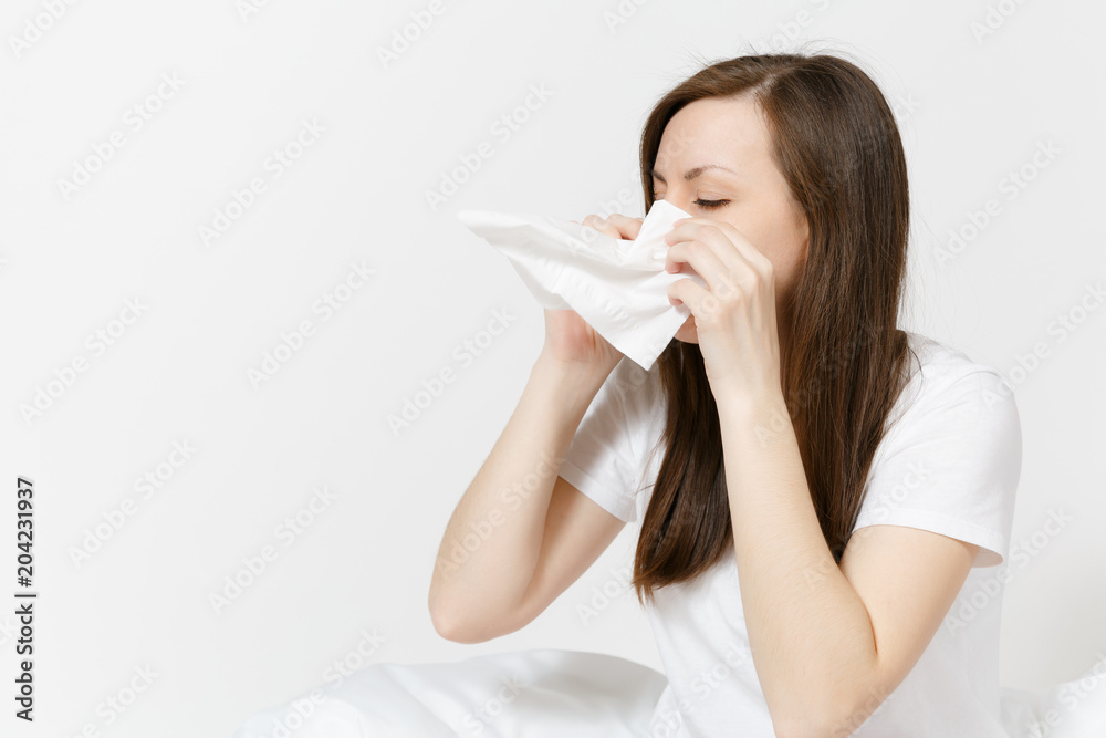 Young sickness tired brunette woman in pajamas sitting on bed isolated on white background. Female feeling bad, blowing her nose into white napkin. Cold in the head. Copy space for advertisement. Young sickness tired brunette woman in pajamas sitting on bed isolated on white background. Female feeling bad, blowing her nose into white napkin. Cold in the head. Copy space for advertisement.