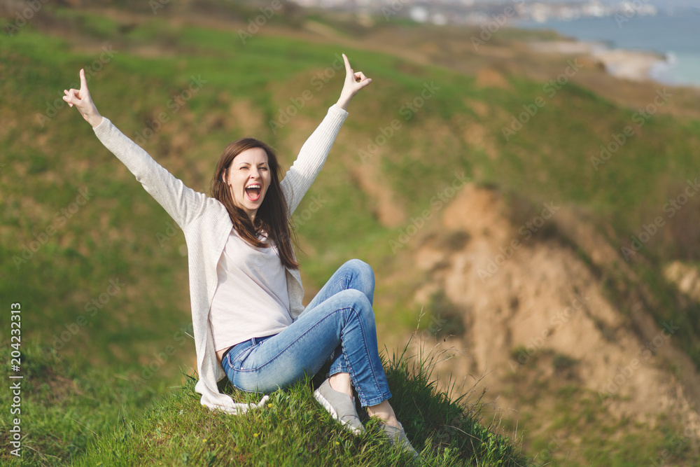 Young fun overjoyed woman in light casual clothes sitting on grass spreading hands screaming showing rock-n-roll sign near water on green field background. Beautiful landscape. Lifestyle concept. Young fun overjoyed woman in light casual clothes sitting on grass spreading hands screaming showing rock-n-roll sign near water on green field background. Beautiful landscape. Lifestyle concept.