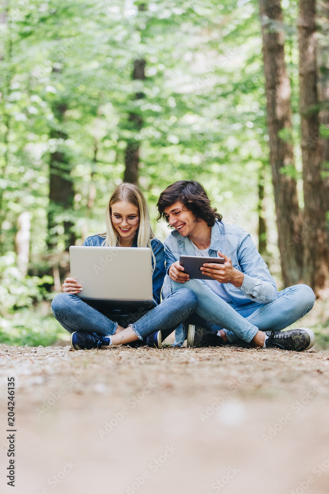 Young couple using laptop and tablet while sitting in forest Young couple using laptop and tablet while sitting in forest