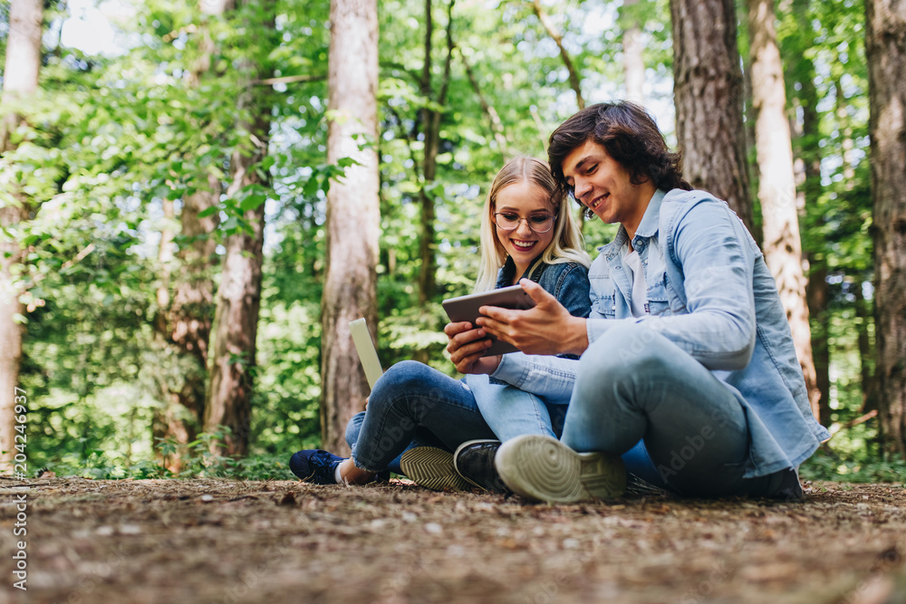 Young couple using laptop and tablet while sitting in forest Young couple using laptop and tablet while sitting in forest