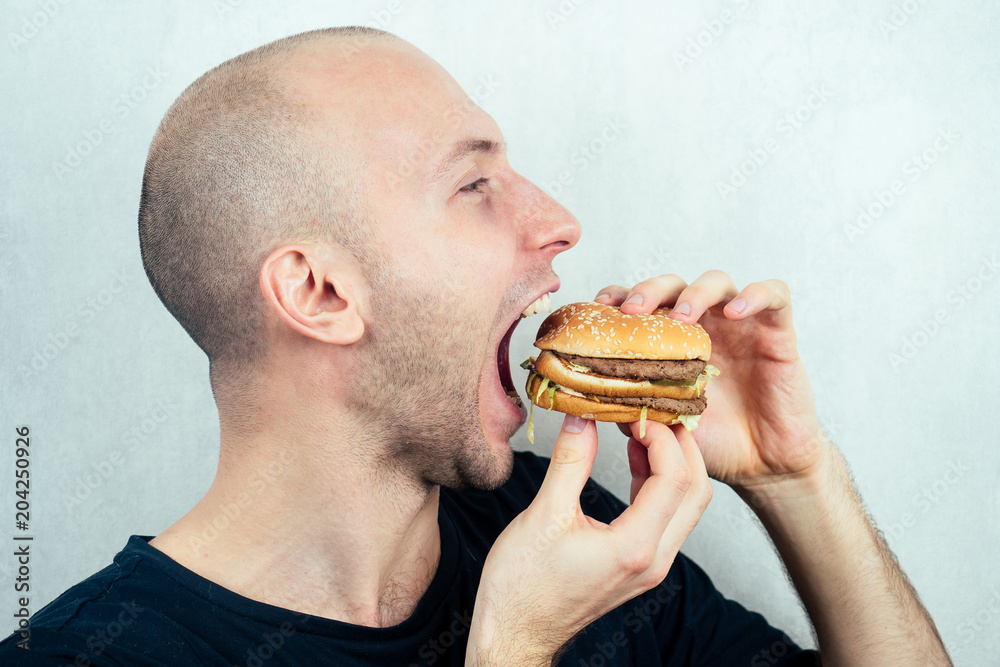 portrait of a young beautiful bald man eats (bites) a high-calorie big burger in his hands. concept of diet, unhealthy and healthy eating portrait of a young beautiful bald man eats (bites) a high-calorie big burger in his hands. concept of diet, unhealthy and healthy eating