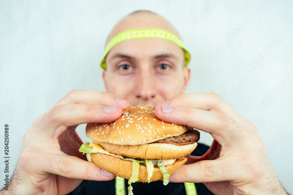 portrait of a young handsome bald hunger man with a measuring tape looking at a high-calorie big burger on a plate. concept of diet, unhealthy and healthy eating portrait of a young handsome bald hunger man with a measuring tape looking at a high-calorie big burger on a plate. concept of diet, unhealthy and healthy eating