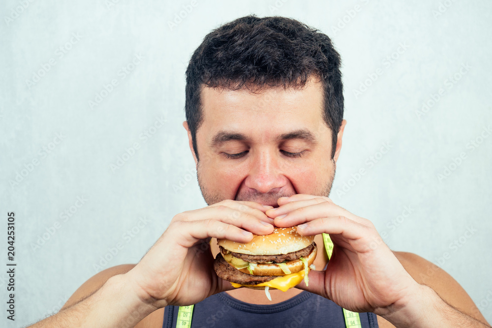 portrait of a young beautiful man eats (bites) a high-calorie big burger in his hands. concept of diet, unhealthy and healthy eating portrait of a young beautiful man eats (bites) a high-calorie big burger in his hands. concept of diet, unhealthy and healthy eating