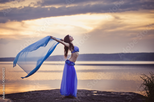 Dancer of bellydance in a blue suit on the beach, against the background of the water. Beautiful nature at sunset. Belly dance. Girl with exotic appearance. Oriental beauty. photo