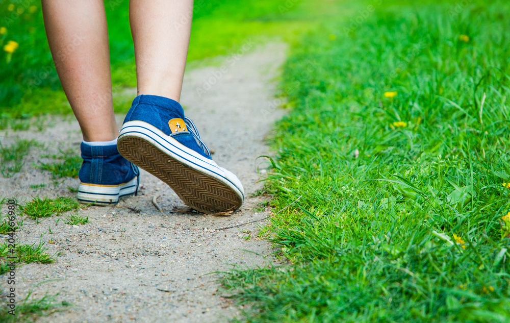Walking women leg in jeans sneaker shoes on the road and green grass