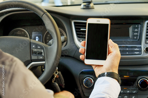 Young man holding blank screen cell phone while driving car. Male driver hand on steering wheel & checking out his smartphone in moving vehicle. Don't text and drive concept. Close up, background.