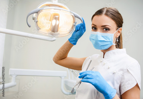 Young female friendly dentist in umiform standing in her office and looking at camera and smiling. Close up potrait of the doctor woman at white background