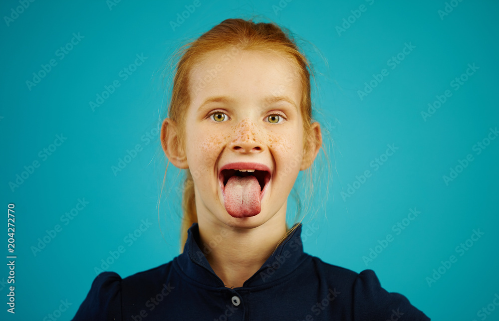 Φωτογραφία Cheerful girl with freckles and red hair opened her mouth wide, stuck out tongue, on isolated blue background