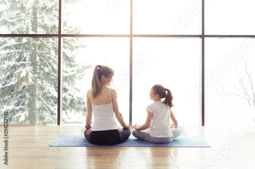 Mother and kid daughter in the gym centre doing yoga poses or stretching fitness exercise near big window with a beautiful scenery with mountains and trees with snow. Healthy family lifestyle concept photo