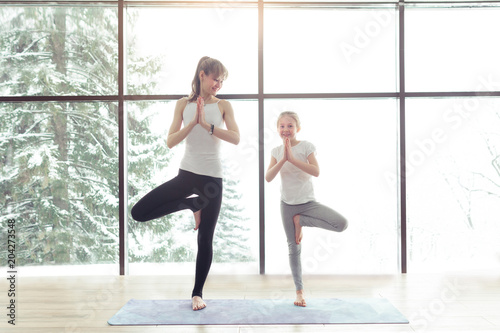 Young beautiful woman and little daughter are doing difficult yoga exercises and smiling while working out at gym with beautiful scenery outside window with sunlight. Healthy fitness yoga concept