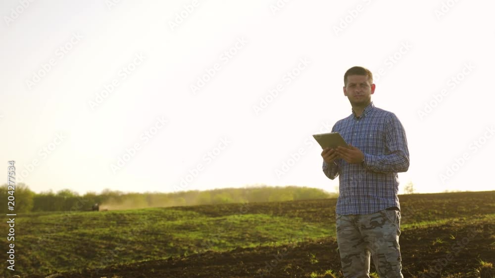 Portrait of a beautiful young man farmer student working in the field with a tractor working in a tablet, happy, dirt in a shirt, the tractor plows a soil ground field. Concept ecology. Smart farming Portrait of a beautiful young man farmer student working in the field with a tractor working in a tablet, happy, dirt in a shirt, the tractor plows a soil ground field. Concept ecology. Smart farming