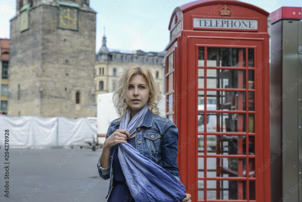 Beautiful blonde model appearance, stands next to a red phone booth.
