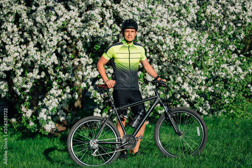 Obraz premium Professional cyclist in helmet and spotswear standing with mountain bicycle near blooming tree. Young rider resting in park after ride.