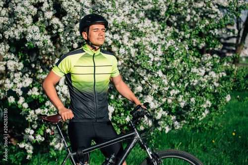 Professional cyclist in helmet and spotswear standing with mountain bicycle near blooming tree. Young rider resting in park after ride. © beatleoff