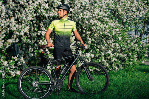 Professional cyclist in helmet and spotswear standing with mountain bicycle near blooming tree. Young rider resting in park after ride. photo